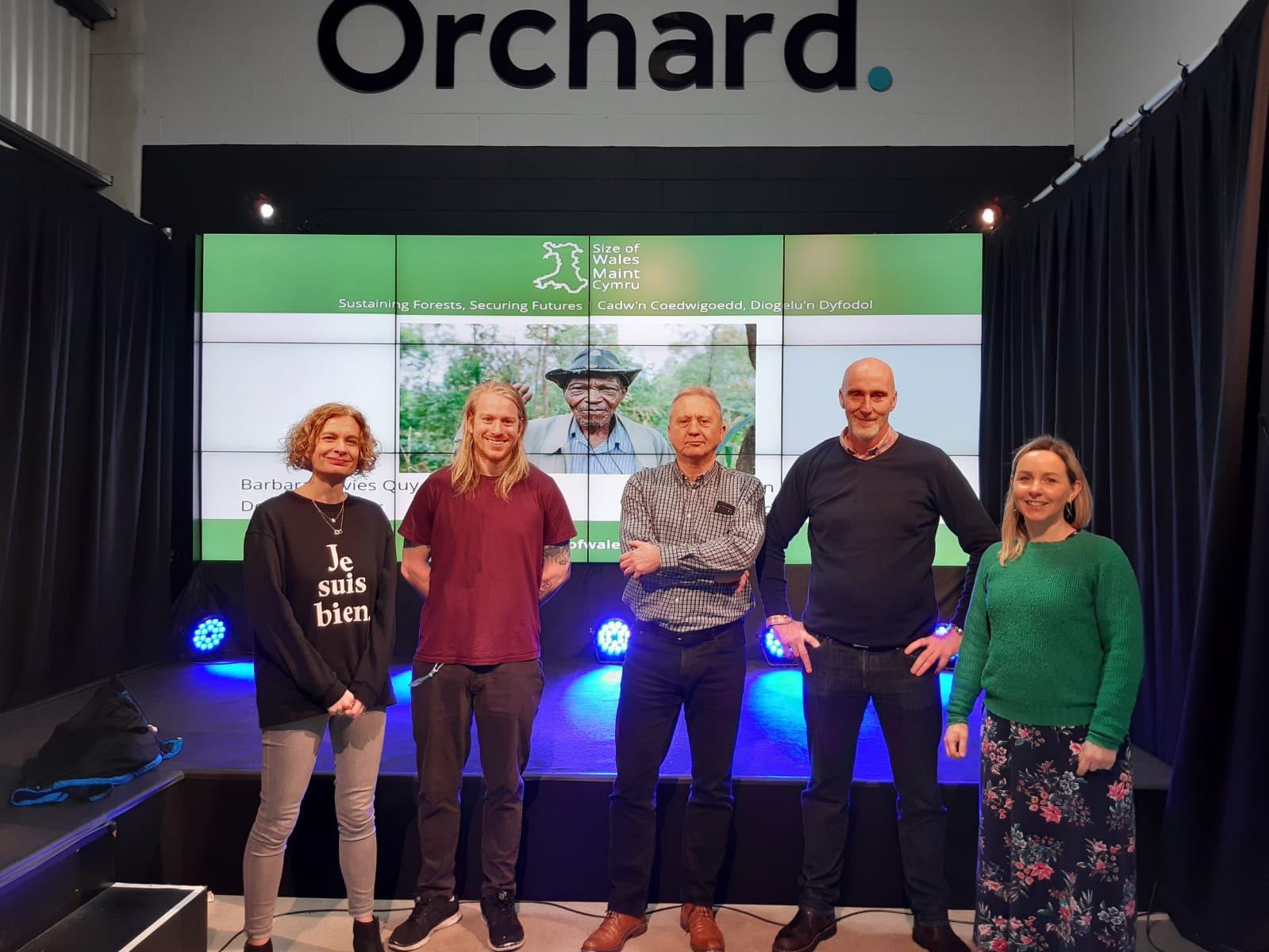 A group of people smiling with the words "Orchard" behind them and the front page of the Size of Wales Website