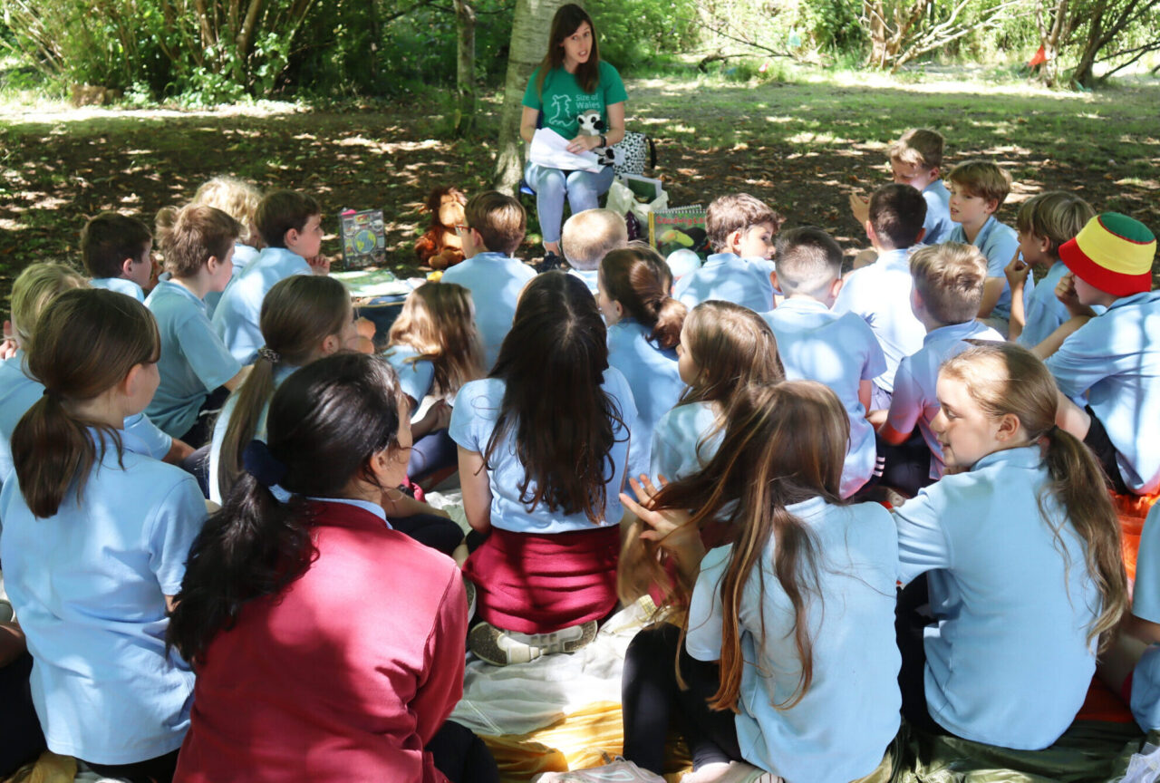 Children sitting under a tree listening to a person in a Size of Wales t-shirt